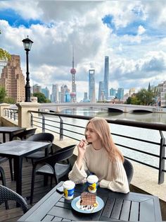 a woman sitting at an outdoor table eating cake and coffee in front of the city skyline