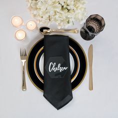 a black and gold place setting with white flowers, silverware, and napkins