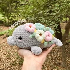 a hand holding a small stuffed animal with flowers on it's head in front of some trees