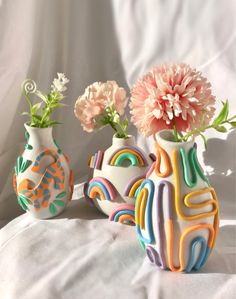 three colorful vases with flowers in them on a white cloth covered surface, one has pink carnations and the other has green leaves