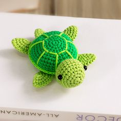 a green crocheted turtle sitting on top of a white table next to a book