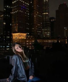 a woman standing in front of a city skyline at night with buildings lit up behind her