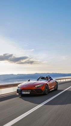 a red sports car driving down the road by the ocean in front of another vehicle