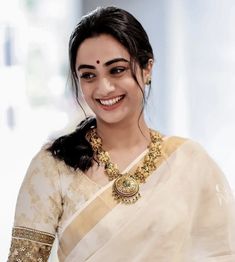 a smiling woman in a white sari with gold jewelry on her neck and shoulder