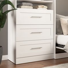 a white dresser sitting next to a bed and a plant on top of a wooden floor