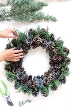 someone is making a pine cone wreath on the table with scissors and greenery next to it