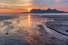 the sun is setting over an ocean with mountains in the distance and sand on the ground