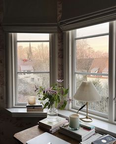 a desk with books and a lamp on top of it next to two large windows