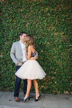 a man and woman standing next to each other in front of a green wall with bushes