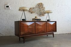 a wooden cabinet with two lamps on top and a wall hanging above it in an empty room