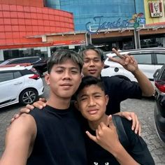 three young men standing next to each other in front of a building with cars parked on the street