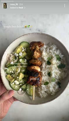 a bowl filled with rice and chicken on top of a table next to a hand holding a skewer