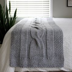 a white bed topped with a gray blanket next to a green potted plant and window