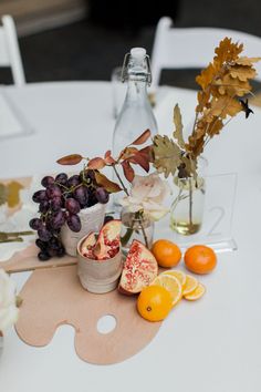 an arrangement of fruit and flowers on a white table cloth with wine bottles, oranges, and grapefruits