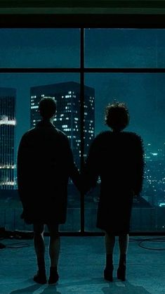 two people standing in front of a window looking out at the city lights and skyscrapers