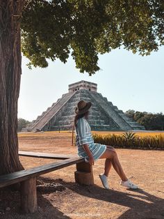 a woman in a hat sitting on a bench near a tree and an ancient structure