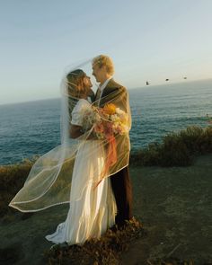 a bride and groom are standing on the cliff by the ocean with their veil blowing in the wind