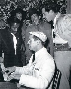 a group of men standing around a man playing a piano in front of another man