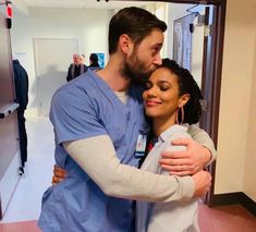 a man and woman hugging each other in a hospital hallway with people walking around the room behind them