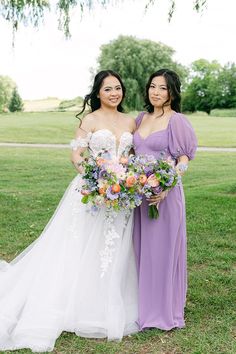 two women in purple dresses standing next to each other holding bouquets and smiling at the camera