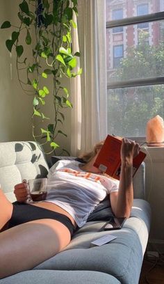 a woman laying on a couch reading a book and holding a drink in her hand