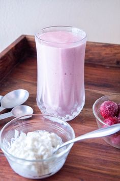 a wooden tray topped with two bowls and spoons filled with yogurt next to a cup of raspberries