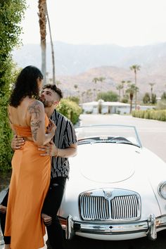 a man and woman standing in front of a white car with palm trees behind them