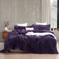 a bed with purple comforter and white pillows in a room next to a window