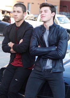 two young men standing next to each other in front of parked cars on a city street