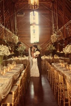 a bride and groom standing in front of their wedding reception tables at the barn with lights strung from the ceiling