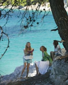 three young women sitting on the rocks by the water looking at something in their hands