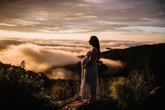 a pregnant woman standing on top of a mountain with clouds in the sky behind her