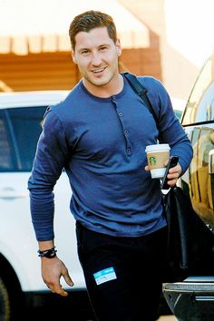 a man in blue shirt holding a coffee cup next to a white car and carrying a black bag