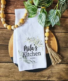 the personalized kitchen towel is next to some green leaves on a wooden plate with a beaded necklace