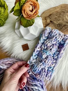 a hand holding a knitted blue and purple blanket next to a white vase filled with flowers