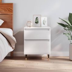 a white nightstand next to a bed with a plant on it and two framed pictures