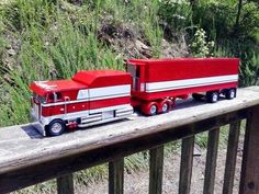 a red and white toy truck on a wooden rail next to some grass, bushes and trees