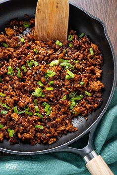 a skillet filled with meat and vegetables on top of a green cloth next to a wooden spatula