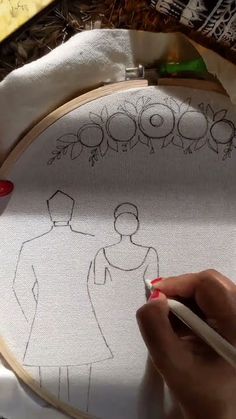 a person is drawing on a piece of fabric with a pencil and scissors in front of an embroidery hoop