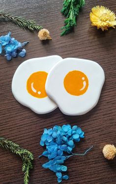 two fried eggs sitting on top of a wooden table next to blue flowers and greenery