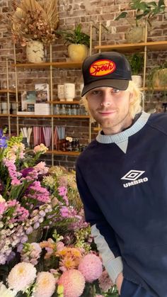 a man standing in front of a bunch of flowers wearing a black hat and blue sweater