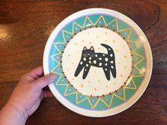 a hand holding a plate with a black cat on it, painted in bright colors