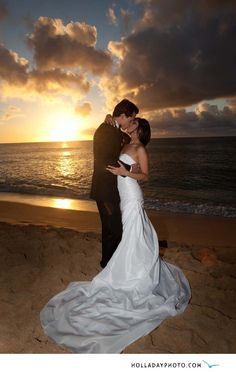 a bride and groom standing on the beach at sunset with their arms around each other