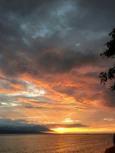 a person sitting on a bench watching the sun set over water with clouds in the sky