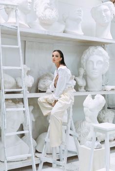 a woman sitting on top of a white chair in front of a shelf filled with sculptures