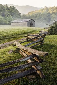 a split rail fence in front of a barn on a foggy day with mountains in the background