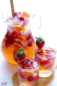 two glasses filled with liquid and strawberries next to pitcher of lemonade on a wooden tray