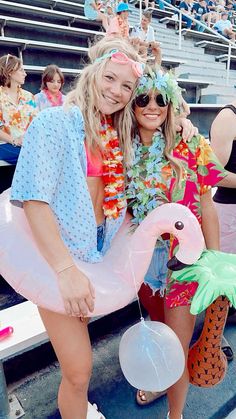 two women in swimsuits pose for a photo at a baseball game with flamingos on the bleachers