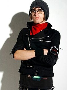 a man with his arms crossed standing in front of a white wall wearing a black jacket and red scarf