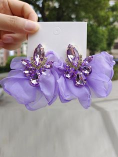 a pair of purple flower clip on earrings with crystal stones in the center, being held up by a person